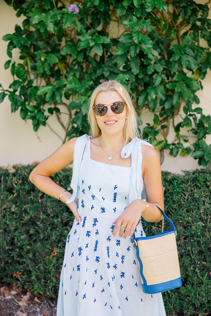 Woman in a floral dress with sunglasses and a rattan straw bag standing in front of greenery.