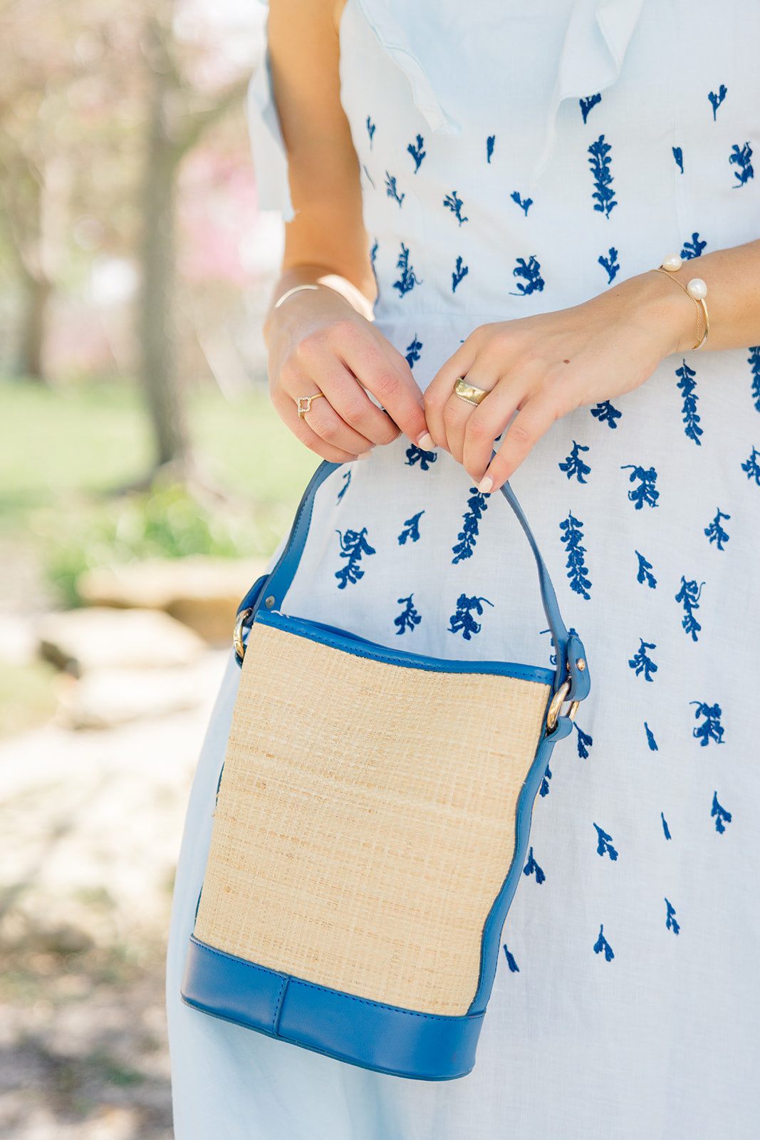 Person wearing a light blue dress with a floral pattern, holding a blue and rattan woven bag outdoors.