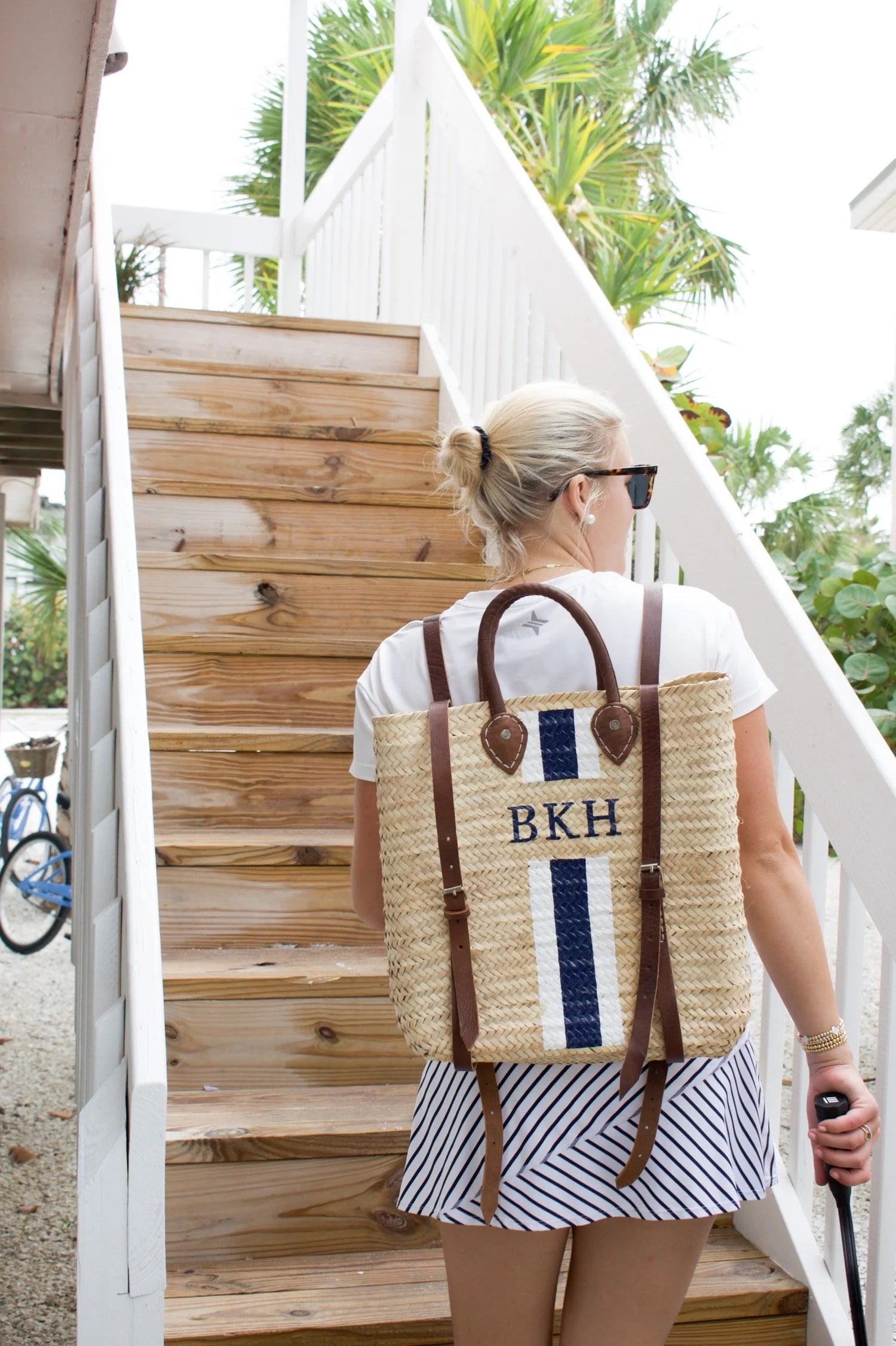 Model walking up stairs in a sunny coastal town carrying the Phebe hand-painted monogrammed straw backpack over both shoulders, paired with a white t-shirt and navy and white stripe skirt.
