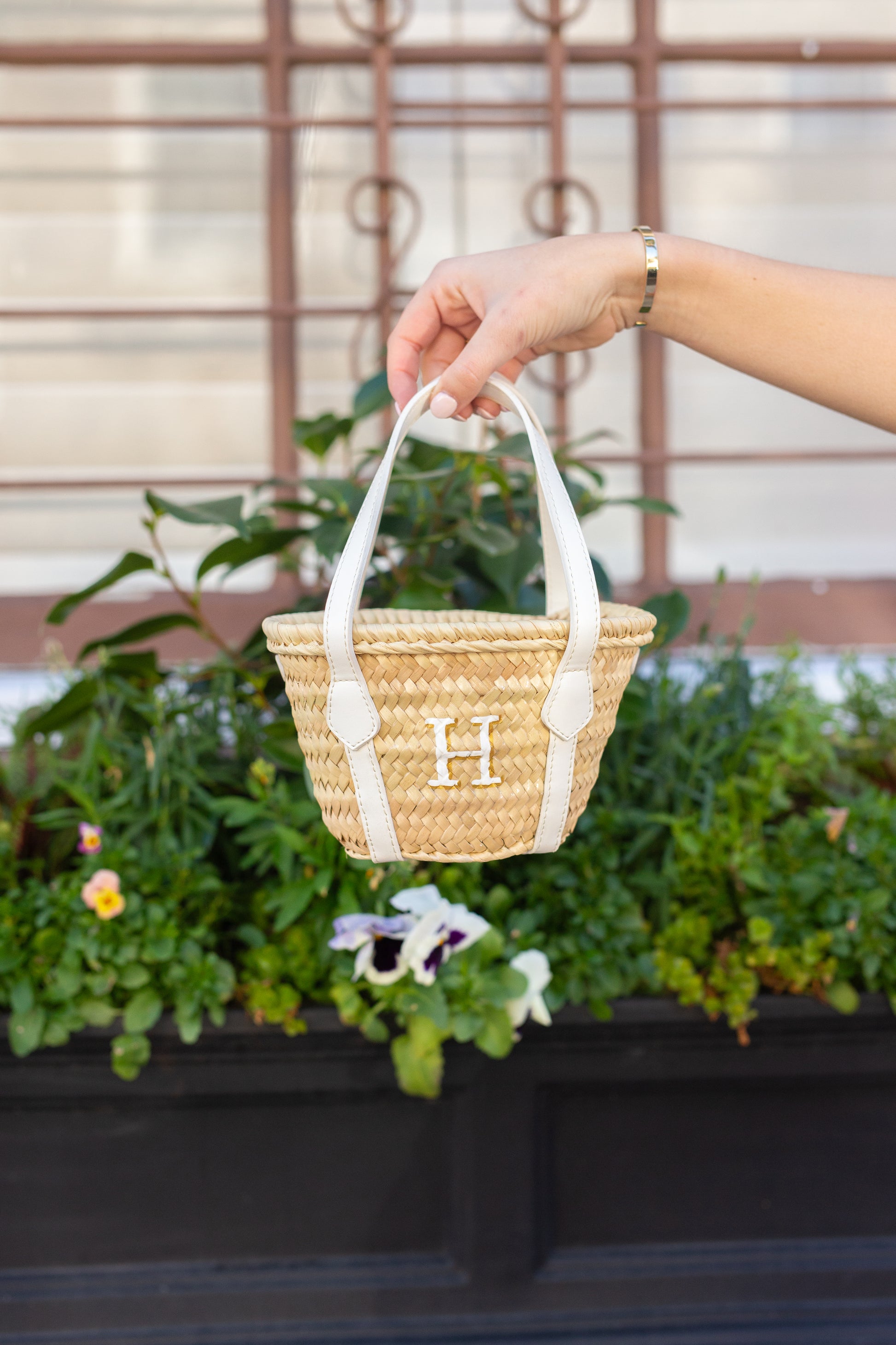 Hand holding a straw handbag with white handles in front of green plants