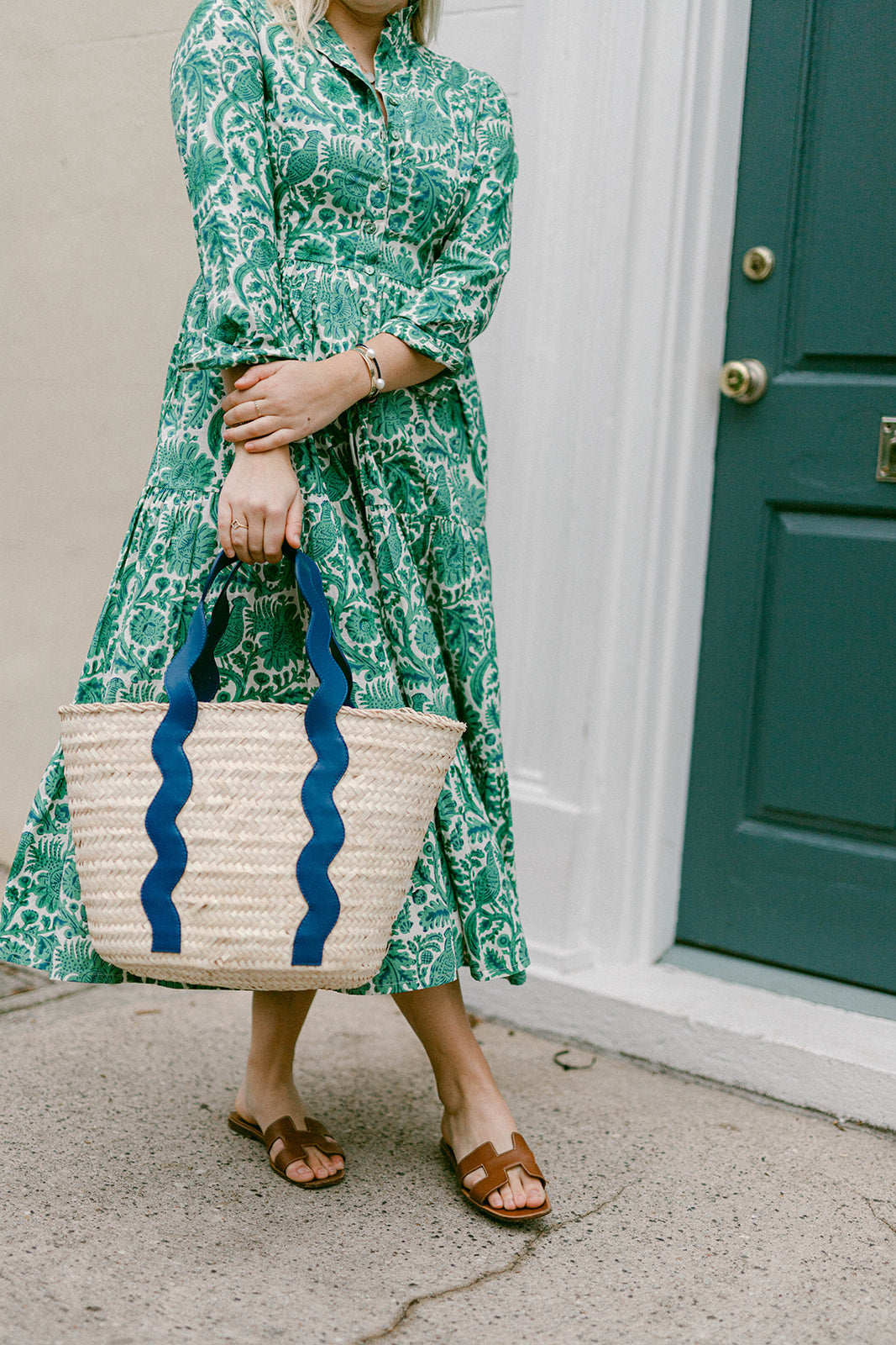 Person wearing a green patterned dress holding a woven bag with blue accents, standing in front of a door.