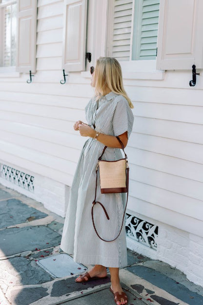 The Boo rattan tote bag styled on a woman in a stripe resort dress, evoking a luxury summer vacation vibe.