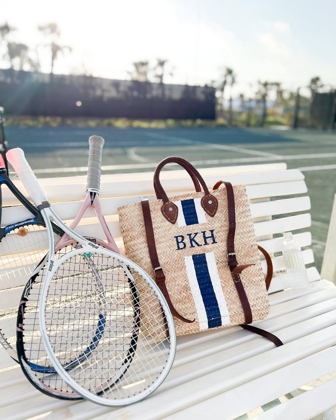 Personalized Phebe backpack showing a custom hand-painted monogram in classic navy and white block lettering while resting on a bench on a tennis court.