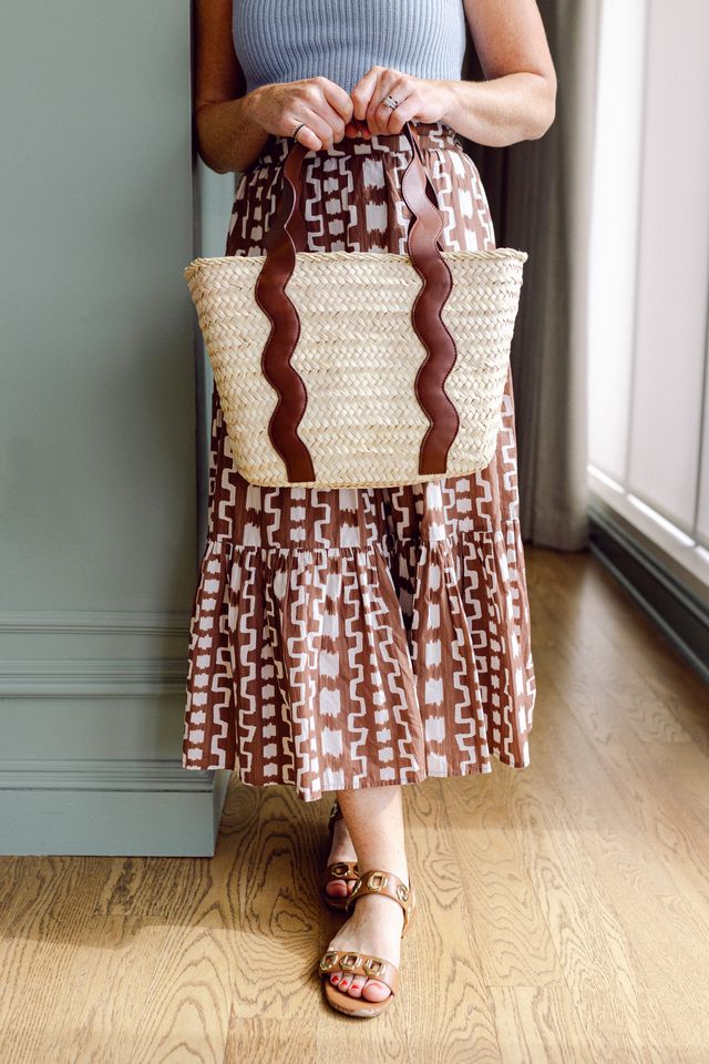 Person wearing a patterned skirt and holding a dark brown woven bag indoors.