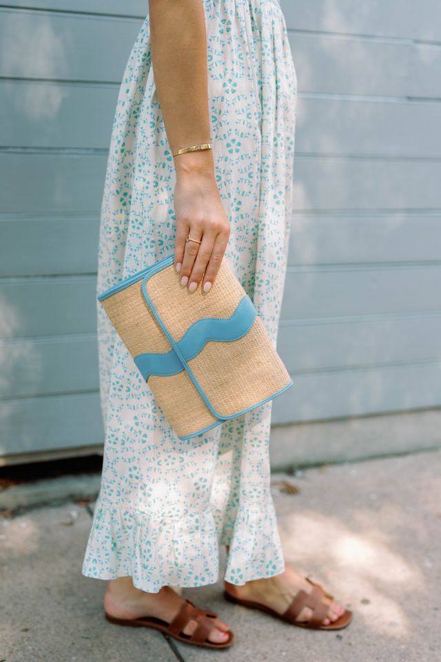 Person wearing a floral dress holding a rattan clutch with french blue accents against a light blue wall.